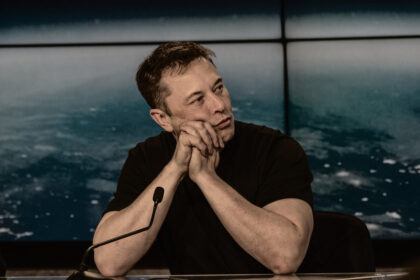 Elon Musk in a black t-shirt sitting at a conference table with a microphone