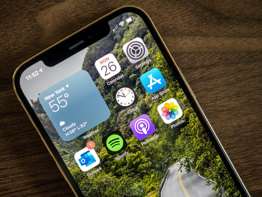 Close-up of iPhone 12 Pro home screen showing apps and weather widget on wooden table